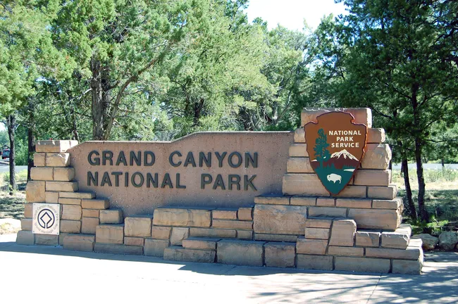 Entrance sign to Grand Canyon National Park surrounded by trees, with the official National Park Service emblem.