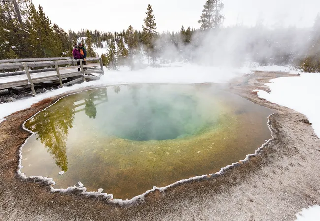 A steaming hot spring with green and yellow water sits in a snowy landscape while two people stand on a wooden boardwalk nearby.