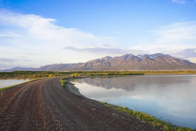 A dirt road curves alongside a reflective body of water at sunset, with the rugged Sangre de Cristo Mountains rising in the distance under a soft, pastel sky.