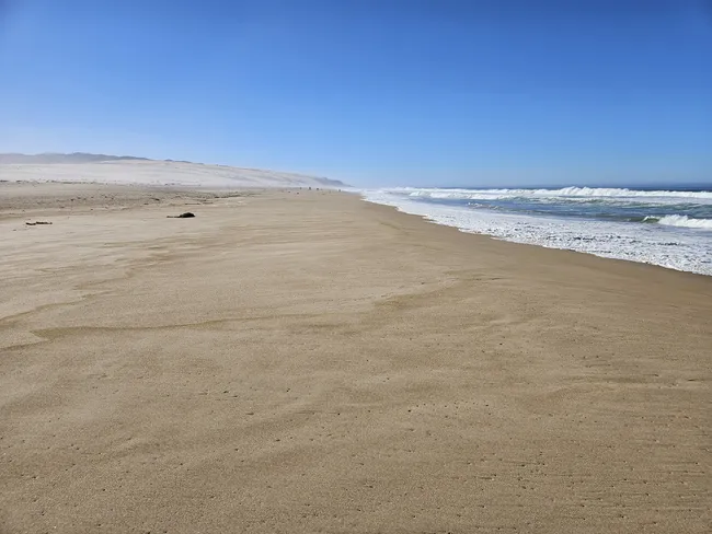 A wide-angle view of a vast, flat sandy beach stretching toward the horizon under a clear, bright blue sky. Gentle white waves crash against the shoreline on the right, and large, pale sand dunes are visible in the far distance on the left.
