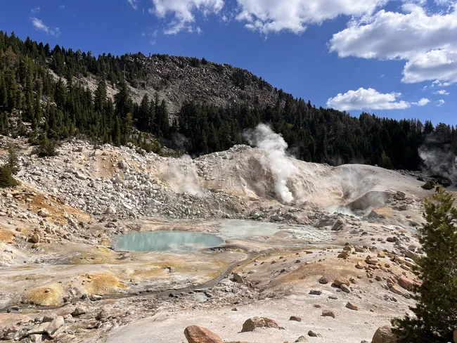 Steam rises from the colorful, rocky terrain of Bumpass Hell as visitors walk along a winding boardwalk.