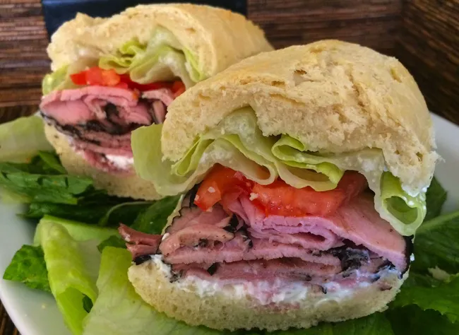 Close-up of a roast beef sandwich with lettuce, tomato, and creamy spread on a sandwich roll, served on a bed of romaine lettuce.