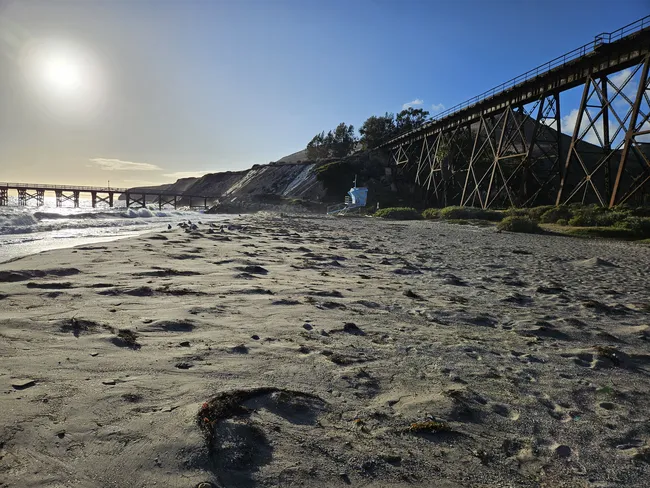 A wide-angle view of a sandy beach at sunset, with a bright sun casting a strong glare over a calm ocean with white crashing waves. A massive, dark metal train trestle rises on the right, stretching toward a steep, light-colored coastal bluff. A small, blue lifeguard stand is perched near the base of the bluff under a clear sky.