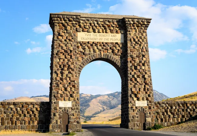 A massive, towering ceremonial archway constructed from rough-hewn, brown-and-grey volcanic stones stands over a paved two-lane road. The structure features a large central arch, two smaller side doors, and a stone tablet at the top engraved with the phrase, "FOR THE BENEFIT AND ENJOYMENT OF THE PEOPLE," set against a backdrop of rolling yellow hills and a bright blue sky with light clouds.