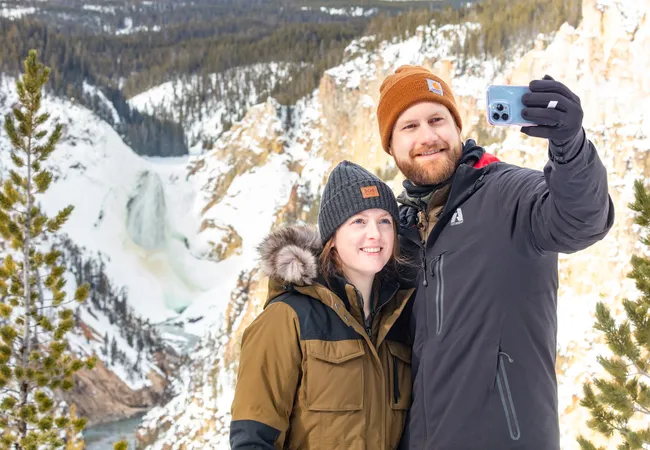 A couple in winter jackets stands close together taking a selfie with a snowy canyon and frozen waterfall behind them.