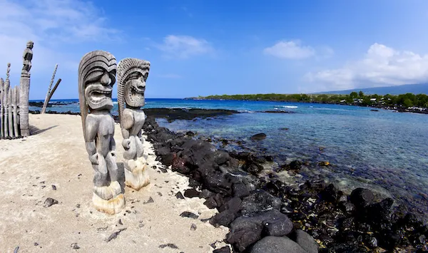 Tiki statues stand on a beach near clear waters, illustrating the importance of cultural respect while visiting Hawaii.
