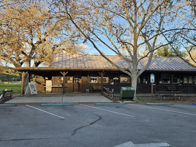 The Pinnacles Camp Store and visitor services building sits beneath large leafless oak trees, with a covered wooden porch, informational signs, and a small parking area illuminated by warm evening light.