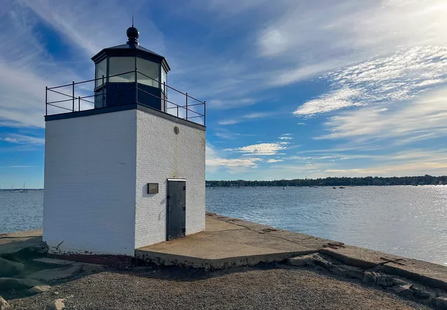 A small square white brick lighthouse with a black top stands at the edge of Derby Wharf, overlooking calm harbor waters under a vibrant blue sky with scattered clouds.