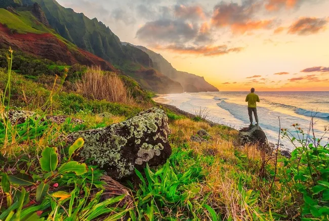 A hiker stands on a coastal cliff overlooking the Na Pali Coast’s rugged mountains and glowing sunset.