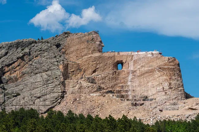Massive mountain carving with a large opening near the top, surrounded by rocky terrain and green pine trees under a blue sky.