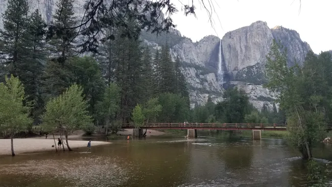 A long wooden footbridge with a simple railing crosses a calm, shallow river where people are wading and standing on a sandy bank. In the background, a very tall, slender waterfall cascades down a massive, sheer grey granite cliff surrounded by dense evergreen trees.