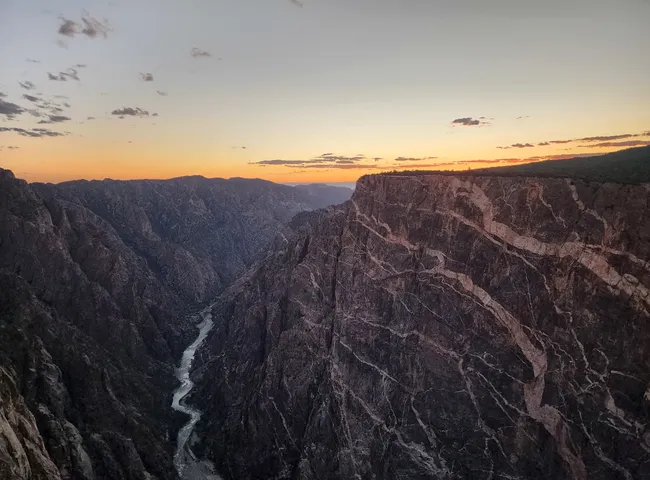 The Gunnison River carves through steep, shadowed canyon walls as a vibrant sunset glows above Black Canyon.