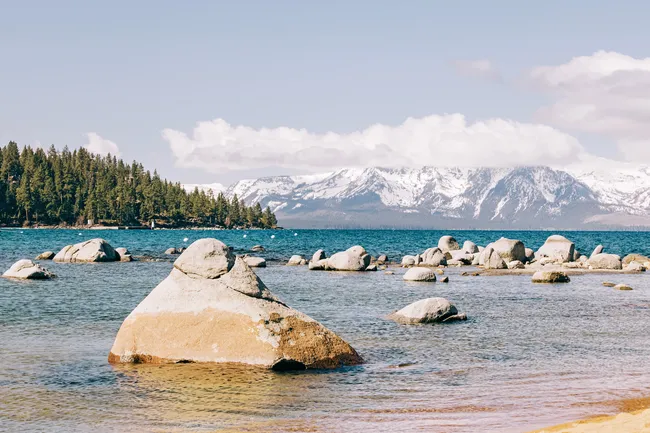Rocky shoreline with calm blue water, distant snow-covered mountains, and a forested peninsula under a partly cloudy sky.