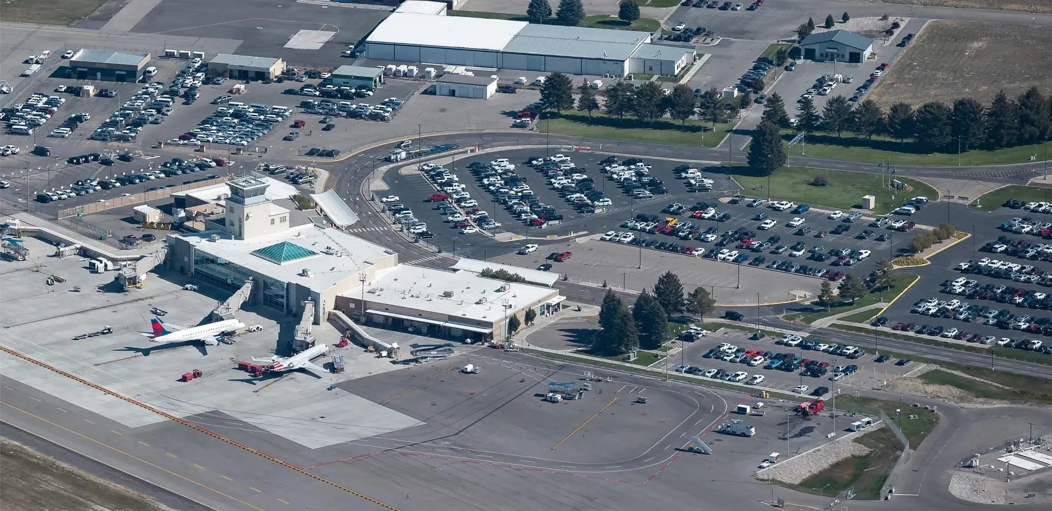 Aerial view of an airport with two airplanes at the gates, terminal buildings, and large surrounding parking lots filled with cars.