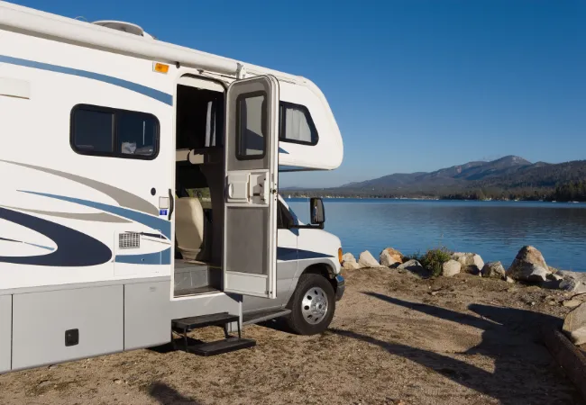 A white RV is parked beside a peaceful lake with mountains in the distance under clear blue skies.