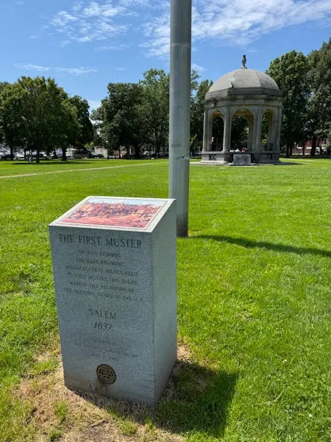 Granite monument in a grassy park in Salem, Massachusetts, marking “The First Muster” of the U.S. National Guard in 1637, with a domed gazebo visible in the background.