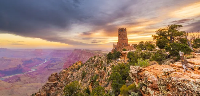 A stone watchtower stands on the canyon’s edge at sunset, overlooking layered cliffs and a winding river far below beneath a dramatic, colorful sky.