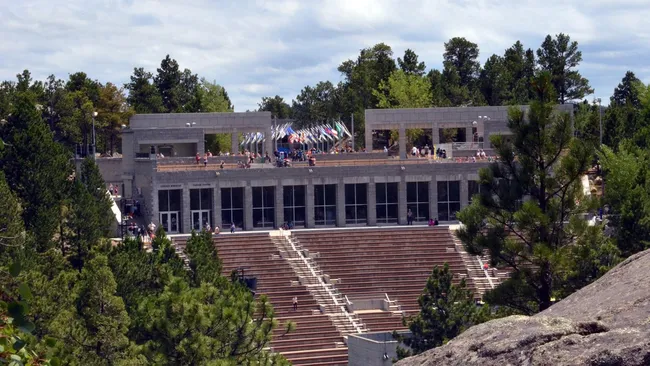 Front view of the Lincoln Borglum Visitor Center and amphitheater at Mount Rushmore, surrounded by trees and American flags on display above