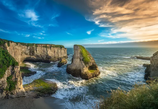 Dramatic coastal cliffs surround a rocky sea stack rising from the ocean, with waves crashing below and golden evening light illuminating the rugged shoreline beneath a colorful sky.