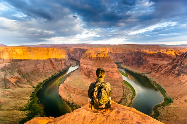 A spectacular high-angle shot from the edge of a steep canyon cliff. A hiker with a backpack sits on the rocky precipice, looking out over the Colorado River as it makes a perfect horseshoe-shaped curve around a massive sandstone formation. The canyon walls are a deep orange and gold, illuminated by the setting sun, while the river below is a dark, emerald green. Dramatic, dark storm clouds fill the sky, with sunlight breaking through to light up the canyon rim.