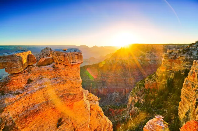 A wide-angle view of a vast canyon at sunrise, with bright golden light and lens flare radiating from the horizon. The sun illuminates the jagged, vertical rock faces and deep, shadowed valleys in warm orange and yellow tones under a clear, transitioning blue sky.