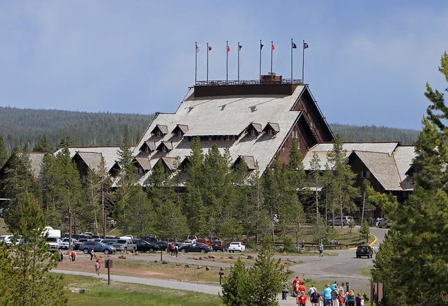 A massive, multi-story wooden lodge with a high-pitched, shingled roof featuring many small dormer windows and several flagpoles at the very top. A busy parking lot and people walking on paths are visible in the foreground.