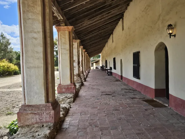 A perspective view looking down a long, shaded outdoor corridor with a floor made of reddish-brown rectangular stone tiles. Thick, cream-colored columns with red accents support a weathered wooden beamed ceiling, casting deep shadows along a white stucco wall with several dark wooden doors and windows.