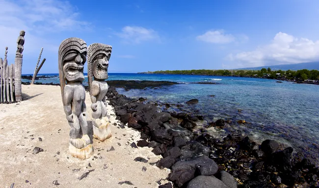 Two traditional Hawaiian wooden statues stand near the rocky shoreline at Puʻuhonua o Hōnaunau National Historical Park.