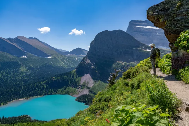 Hiker stands on Grinnell Glacier Trail overlooking a turquoise lake and rugged mountain peaks in Glacier National Park.