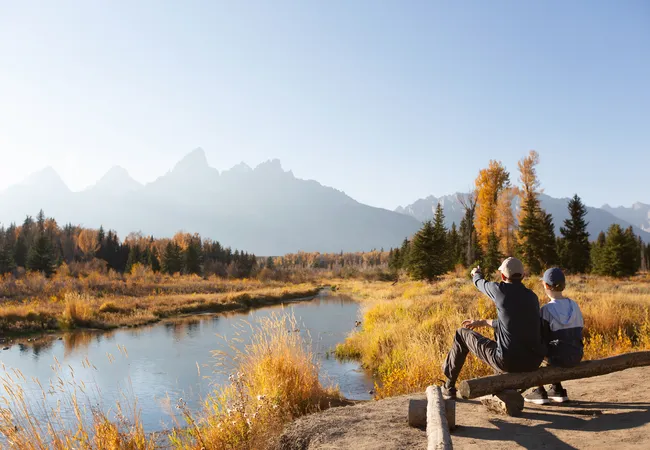 Two people sitting on a log by a stream, looking at autumn scenery of mountains and trees, illustrating a kid-friendly hiking trail experience.