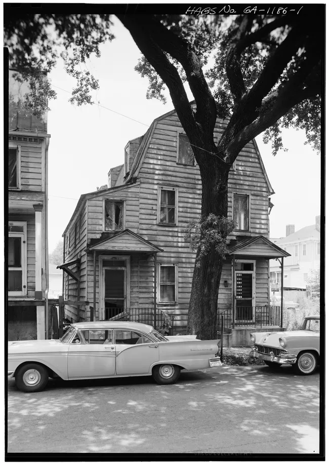 Historic wooden house with a large tree and vintage cars parked in front, located in a residential area.