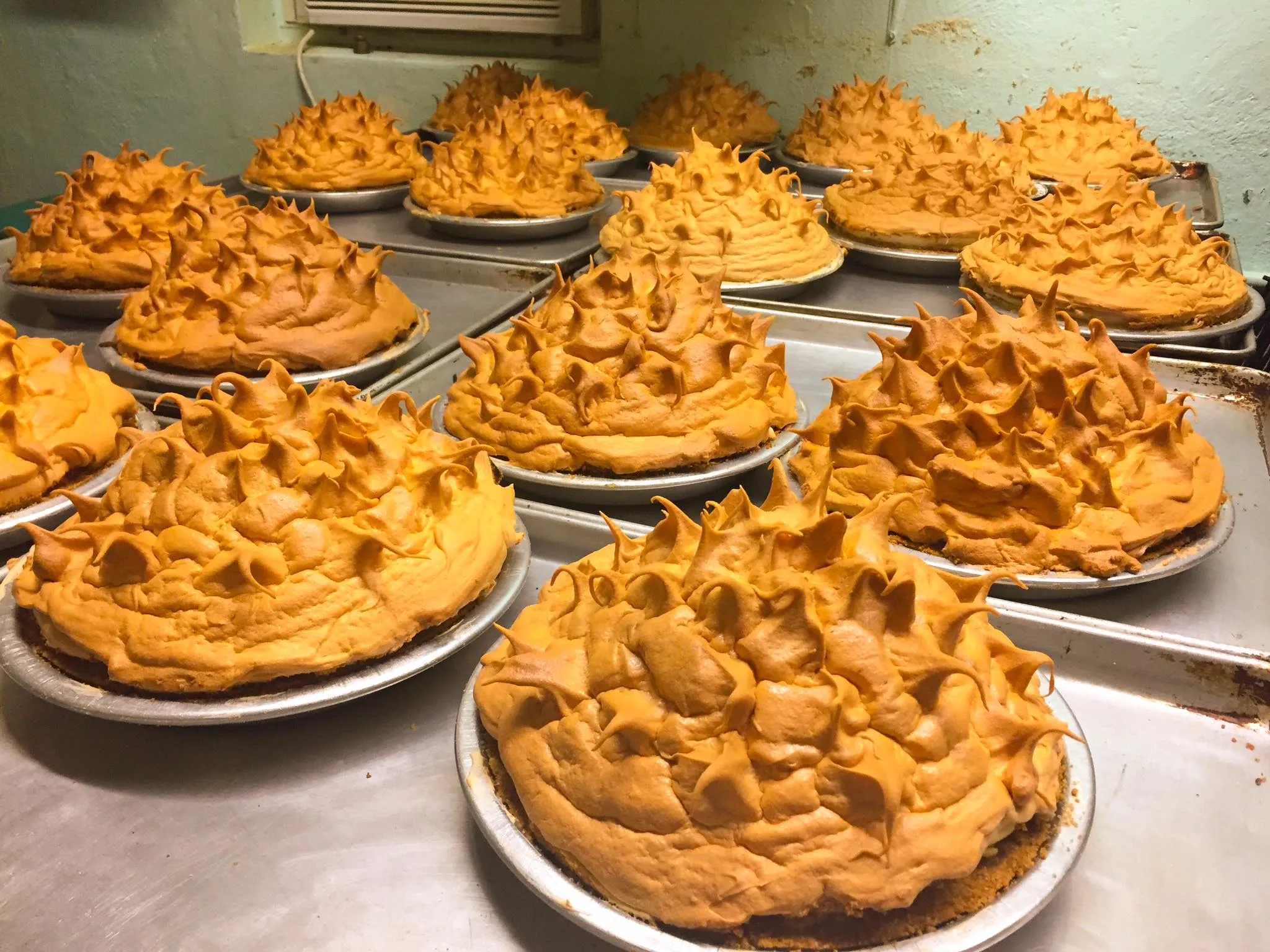 A high-angle, wide shot of dozens of freshly baked key lime pies cooling on a metal kitchen counter. Each pie is topped with a massive, toasted golden-brown meringue that has been peaked into high, jagged points. The pies sit in silver tin pans on flat baking sheets, stretching into the background of a professional kitchen.