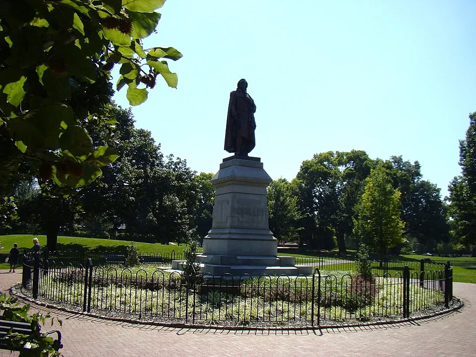 A bronze statue of Friedrich Schiller, the German poet and playwright, standing atop a tall stone pedestal inscribed with "Schiller," set within a circular garden bed enclosed by a low black iron fence. The monument is surrounded by a red brick pathway, park benches, and lush green lawns shaded by large mature trees. A person is visible walking in the background to the left. The photo is taken on a bright, clear sunny day in Schiller Park, German Village, Columbus, Ohio.