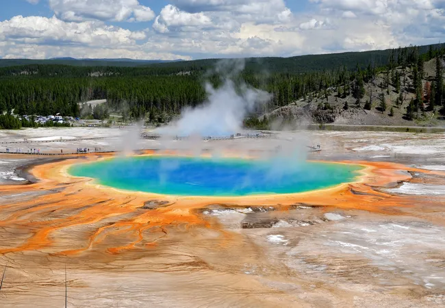 A high-angle view of a massive circular geothermal spring with a deep blue center, surrounded by concentric rings of bright teal, green, and vivid orange mineral deposits. Plumes of white steam rise from the water's surface, set against a backdrop of dark green forests and a cloudy sky.