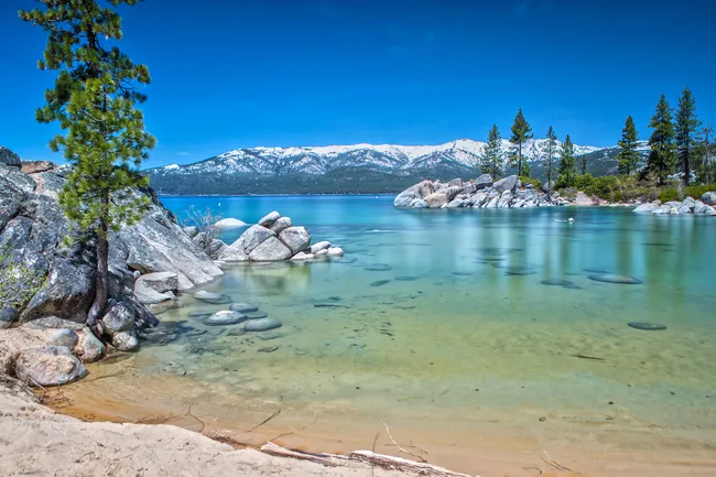 Clear turquoise lake with sandy shore, rocky edges, pine trees, and snow-capped mountains in the background under a bright sky.