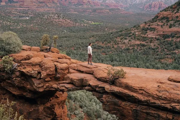 A hiker stands on a narrow red rock arch overlooking a vast desert valley filled with green vegetation and rugged cliffs.