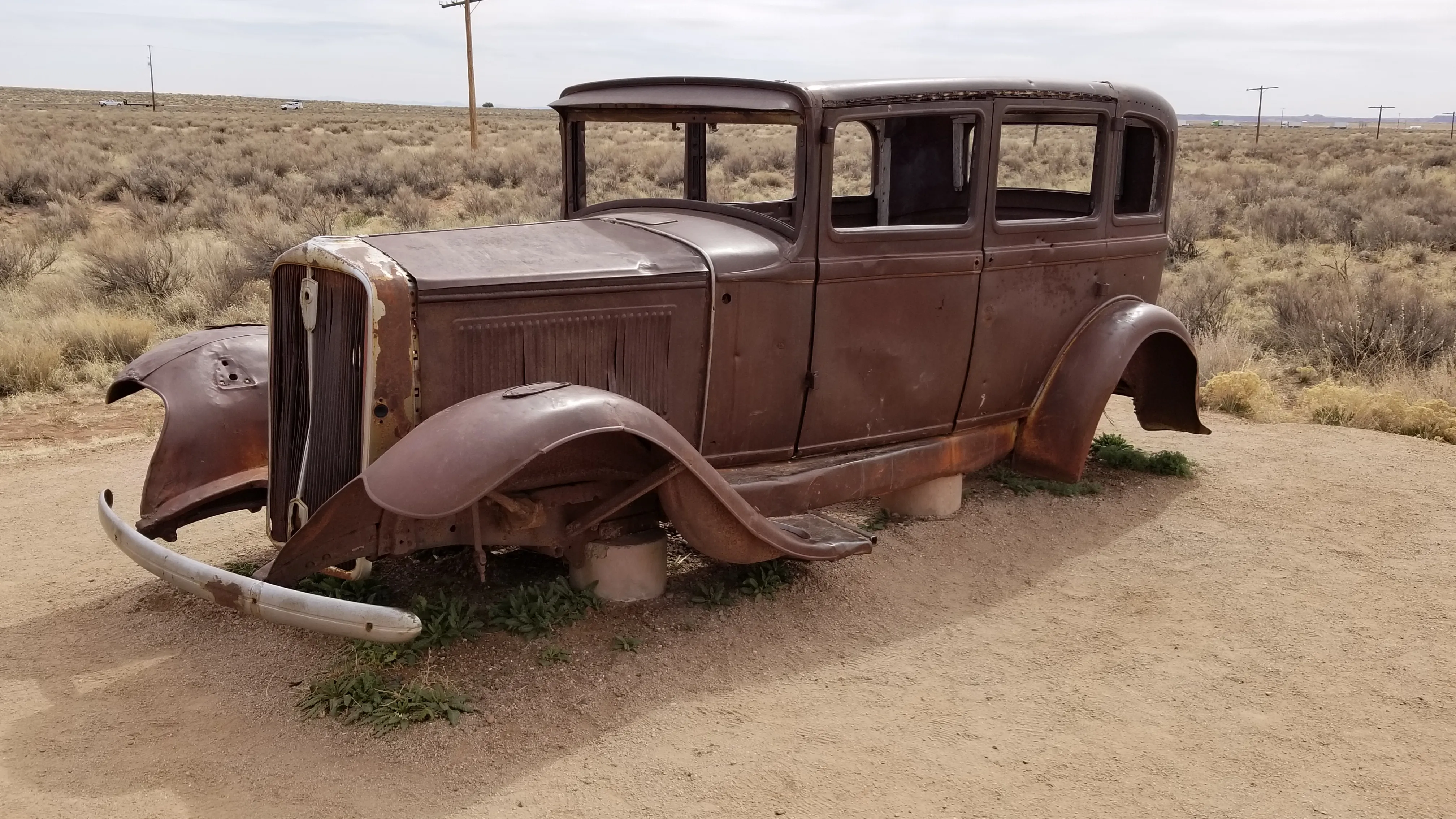 A wide, side-angle shot of a rusted, vintage 1930s-era car frame sitting in the middle of a vast, dry desert landscape. The car is missing its wheels, windows, and engine components, resting on stone blocks over light-colored dirt. In the background, a flat horizon of dry scrubland stretches under a cloudy sky, with several utility poles fading into the distance.