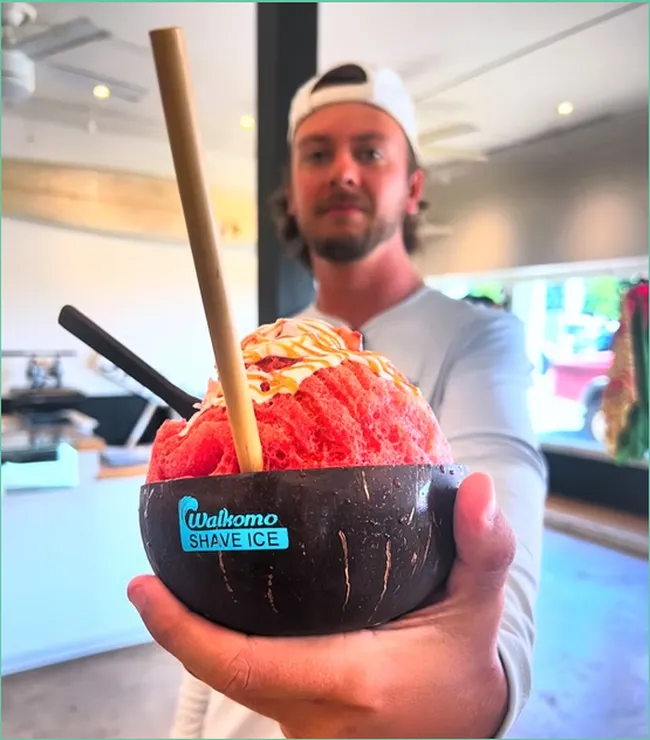 Man holding a colorful bowl of strawberry-flavored shave ice topped with syrup from Ululani’s Shave Ice.