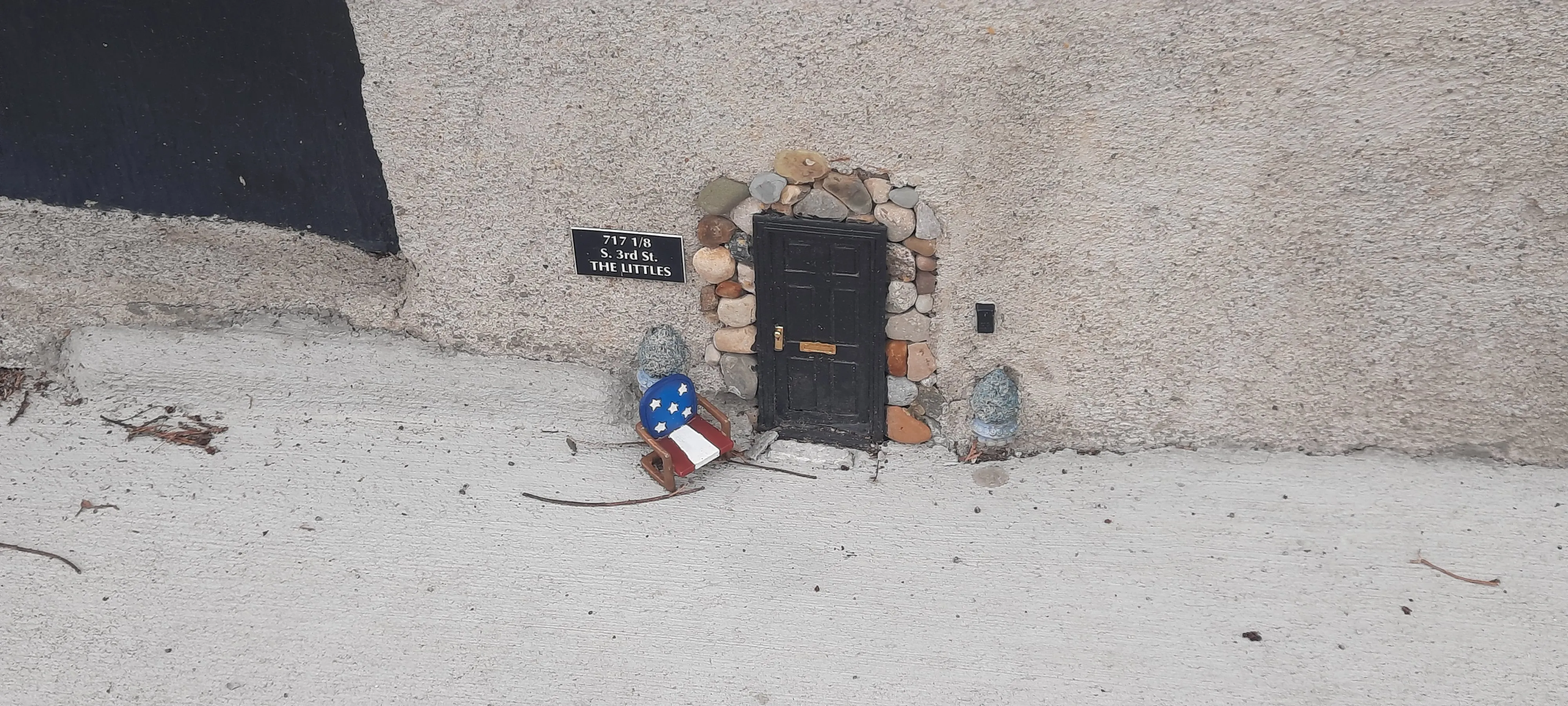 A tiny miniature black door built into the base of a textured concrete wall, framed by small river rocks. A small address plaque to the left reads "717 1/8 S. 3rd St. — THE LITTLES." In front of the door sits a miniature rocking chair painted in American flag colors (red, white, and blue with white stars), along with two small stone figurines flanking the entrance. The whimsical installation is set at ground level on a concrete sidewalk.