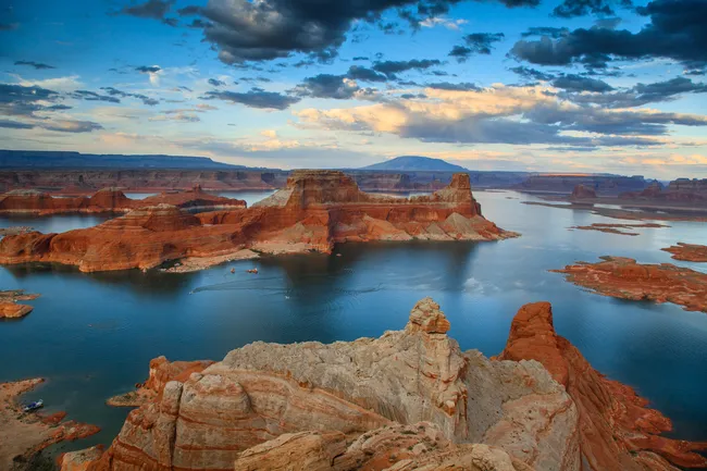 Red rock formations rise from a calm blue lake, with layered cliffs and distant mountains beneath a sky filled with soft, dramatic clouds.