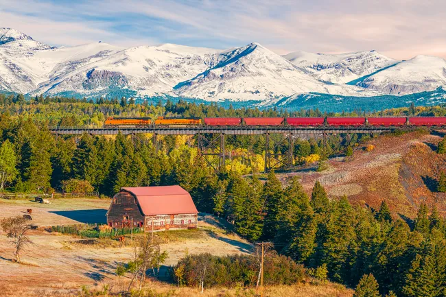 Freight train crosses a high railway bridge with snowy mountains and a red-roofed barn in East Glacier Park Village.