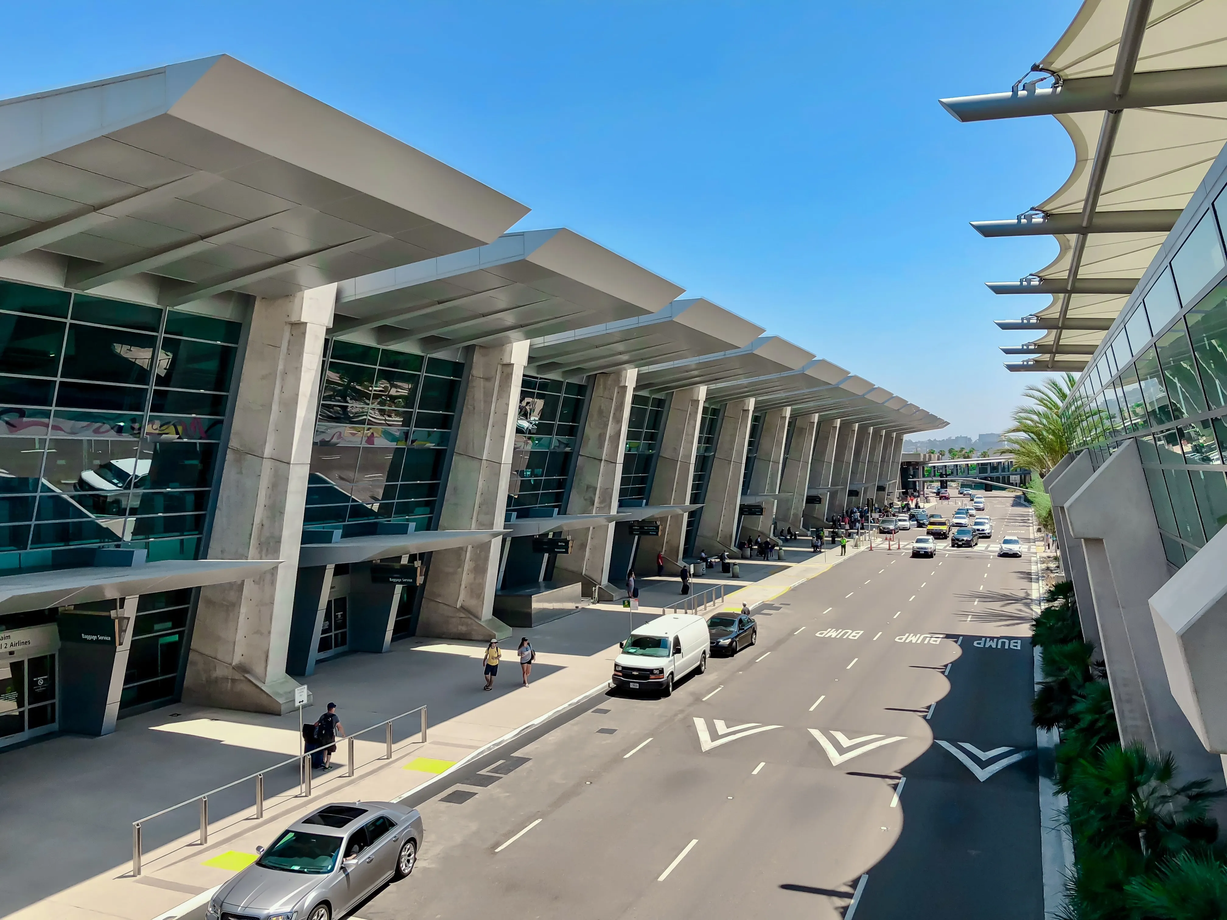 A high-angle view of a modern airport terminal featuring a long, slanted glass facade supported by several massive concrete pillars. A multi-lane roadway with light traffic and "BUMP" markings stretches past the building under a clear blue sky, with a few travelers walking on the wide sidewalk near parked cars.