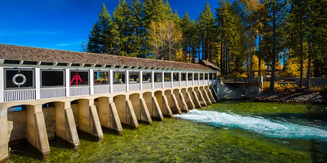 A long covered bridge-style dam in Tahoe City spans clear green water, surrounded by tall pine trees and autumn colors.