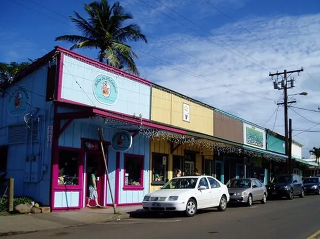 Colorful storefronts in the small town of Paia, Maui, with parked cars and a palm tree overhead.