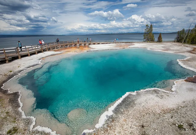 A high-angle view of a deep, crystal-clear turquoise pool with a bright white, jagged mineral rim. A wooden boardwalk with several people on it curves along the edge of the pool, separating it from a vast, calm lake in the background under a sky filled with dramatic white and grey clouds.
