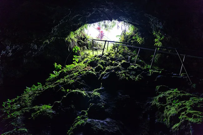 Moss-covered cave interior with a metal railing, looking out toward bright tropical greenery at the cave entrance.