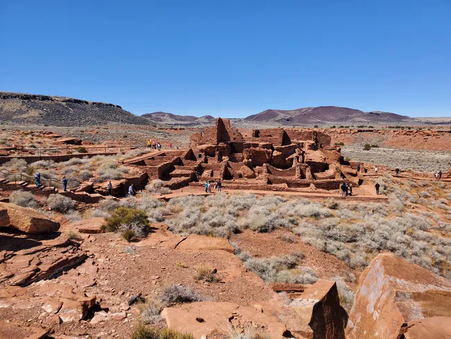 Ruins of a large red sandstone pueblo structure at Wupatki National Monument, surrounded by desert and distant hills.