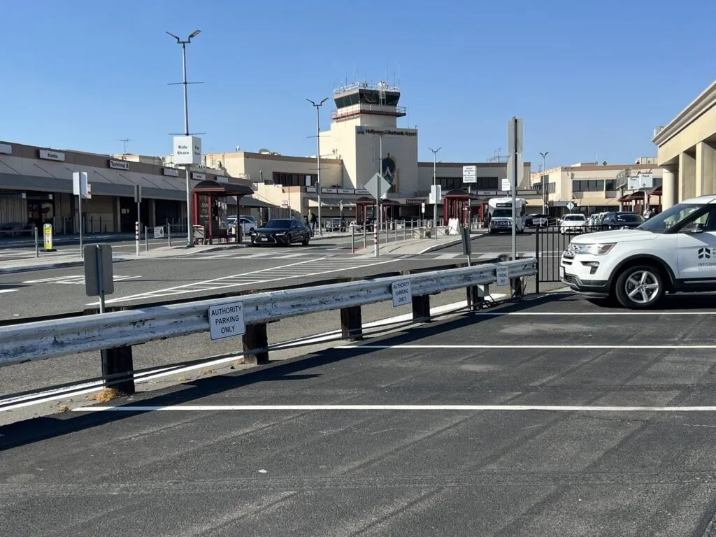 A ground-level view of an airport terminal and control tower under a clear blue sky. In the foreground, a paved parking lot with a "Authority Parking Only" sign on a metal guardrail sits next to a white SUV. Across the roadway, the main terminal building is visible with signage for ride-sharing and various airlines.