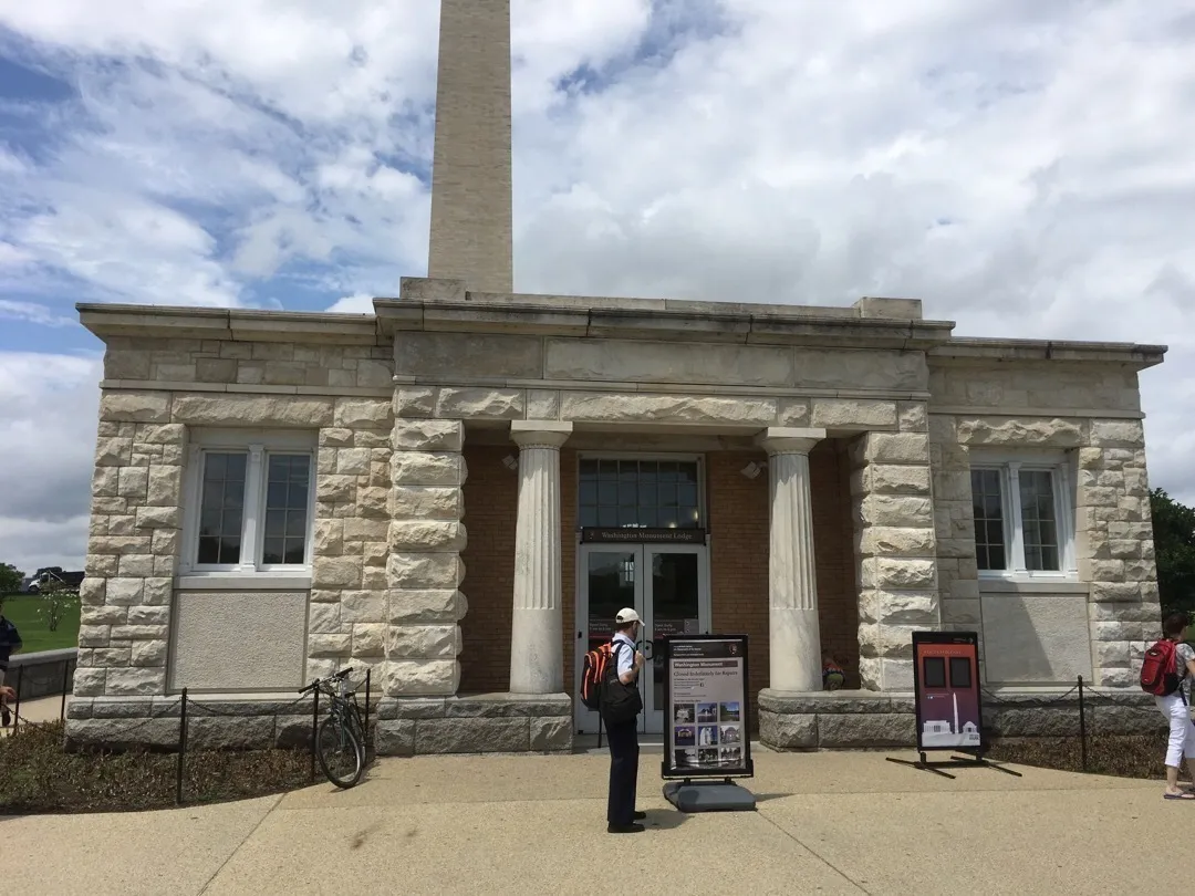 The Washington Monument Lodge, a small neoclassical stone building with white limestone block walls and four columned columns flanking the entrance, situated at the base of the Washington Monument in Washington, D.C. The tall white obelisk of the monument rises prominently behind and above the lodge. A person wearing a backpack and cap stands in front reading an informational sign, with a bicycle parked nearby. Additional visitors are visible on either side. The sky is partly cloudy.
