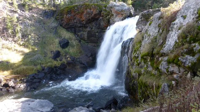 A small waterfall cascades over mossy rock cliffs into a clear pool surrounded by forested terrain.